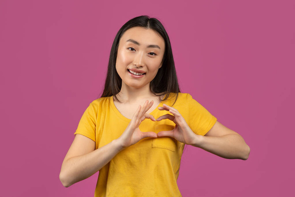 Szczęśliwy Azji lady pokazując gest serca z rąk w pobliżu klatki piersiowej, co znak miłości, pozowanie nad fioletowym tle studio - Zdjęcie, obraz