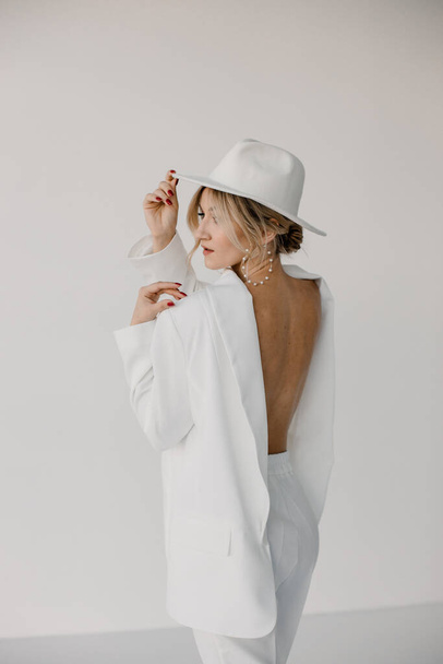 Confident young blond woman smiling, looking at camera isolated on white background. Studio portrait of successful friendly female in white suit and hat, posing over white wall. - Фото, изображение