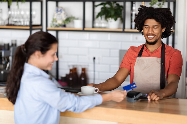 Happy millennial african american curly male waiter in apron gives fresh coffee to client - Fotó, kép