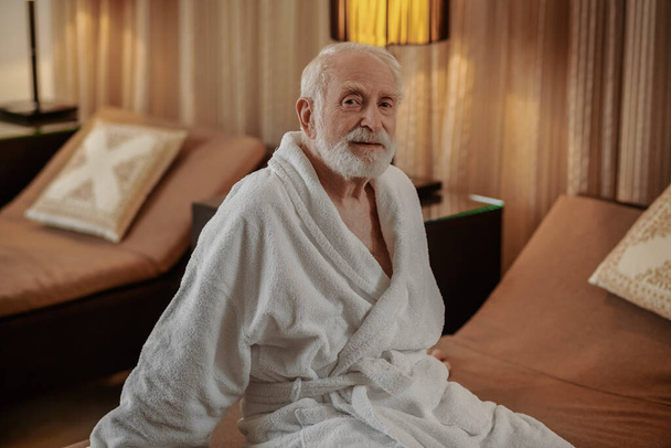 Сіроволоса бородата людина в білому халаті в готельному номері
 - Фото, зображення
