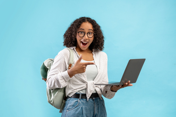 Emocjonalne młoda czarna kobieta z plecakiem wskazując na ekranie laptopa w podnieceniu nad niebieskim tle studio - Zdjęcie, obraz