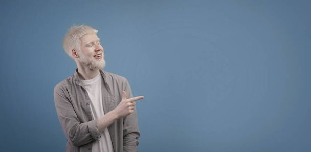 Schauen Sie zur Seite. Junger Albino-Mann zeigt mit dem Finger zur Seite und zeigt Kopierfläche für Werbetext, blauer Hintergrund - Foto, Bild