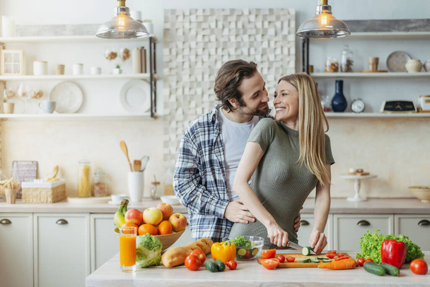Satisfied young european male with stubble hugs female, prepares food at table with organic vegetables in kitchen - Photo, Image