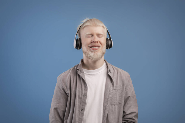 Задоволений спокійний альбіно-хлопець в навушниках насолоджується улюбленою музикою з закритими очима, що стоять над синім тлом
 - Фото, зображення