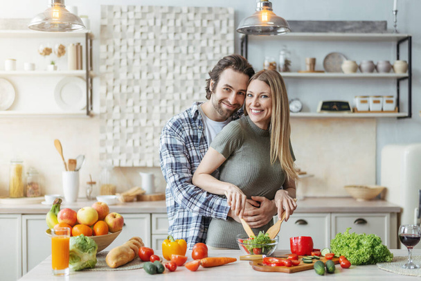 Cheerful young caucasian lady cooking salad, guy with stubble hugs woman at table with organic vegetables - Foto, afbeelding