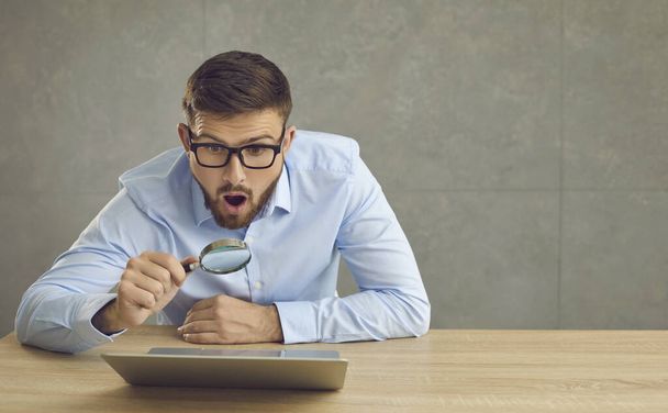 Man with a surprised expression looks at the laptop screen through a magnifying glass. - 写真・画像