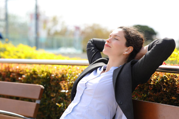 Relaxed businesswoman resting with hands on head sitting on a bench in a park - Photo, image