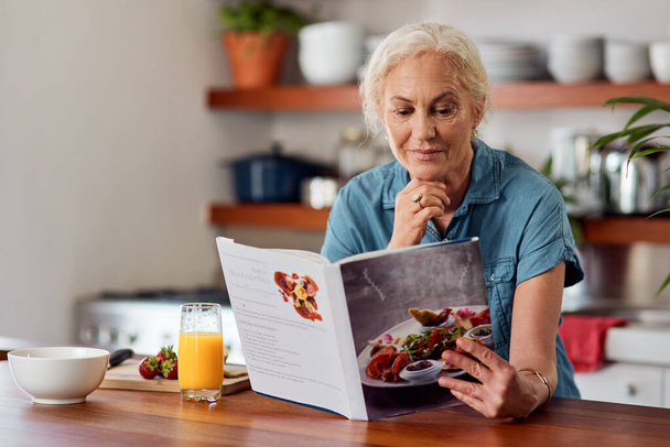 Der gesunde Weg ist der intelligente. Aufnahme einer reifen Frau, die ein Buch liest, während sie zu Hause das Frühstück zubereitet. - Foto, Bild