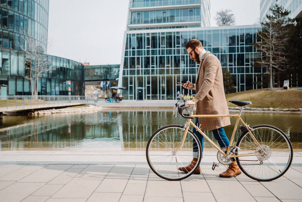 Young hipster businessman walking with bicycle. Using smartphone. Eco friendly transport - Fotó, kép