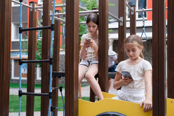 two charming teenage girls in identical clothes with smartphones outside. kids surf the net. smartphone addiction. children in social networks. texting, playing, communicate. - Photo, Image