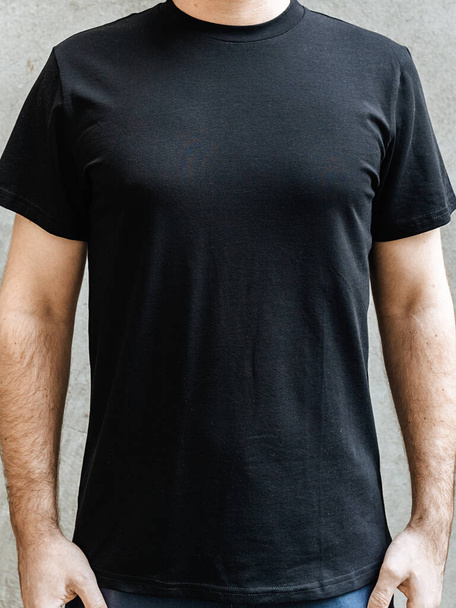 Young man in an empty black casual t-shirt. Front view close up on a background of light gray concrete wall. Design and layout of men's t-shirt for printing. - Foto, Imagem