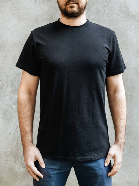 Young man with a beard in an empty black casual t-shirt. Front view on a background of light gray concrete wall. Design and layout of men's t-shirt for printing. - Foto, imagen