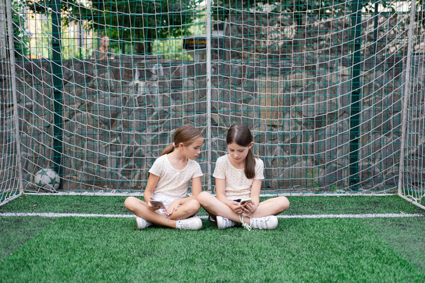 two pretty girls in identical clothes using phone on sports ground or athletic field. kids are surfing in internet. smartphone addiction. advanced, modern children in social networks. - Фото, изображение