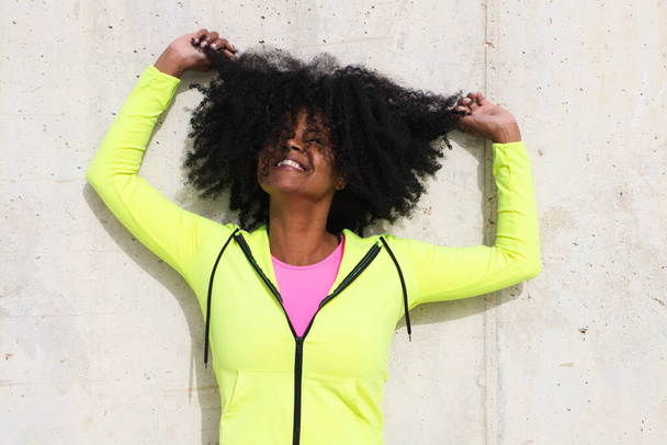 Beautiful young Afro-American woman in bright green and pink sportswear on a grey concrete wall texture background. Woman makes different expressions. Laughing, serious, happy, sad, thinking, hair. - Photo, Image