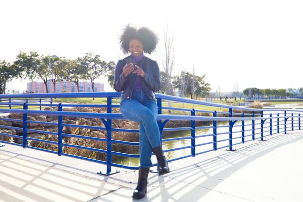 Νεαρή, όμορφη Αφρο-Αμερικανίδα γυναίκα με κινητό τηλέφωνο να ψωνίζει online, τσεκάροντας τα email και τα κοινωνικά δίκτυα στο πάρκο. Η γυναίκα φοράει καθημερινά και άνετα ρούχα και είναι ευτυχισμένη. - Φωτογραφία, εικόνα