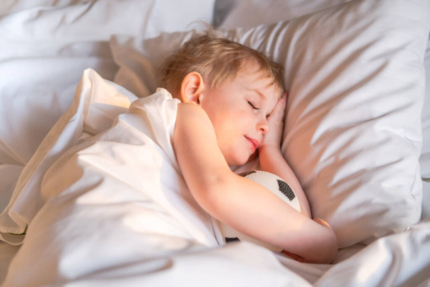 Toddler boy sleeping with a soccer ball on a white bed linen. - Foto, imagen