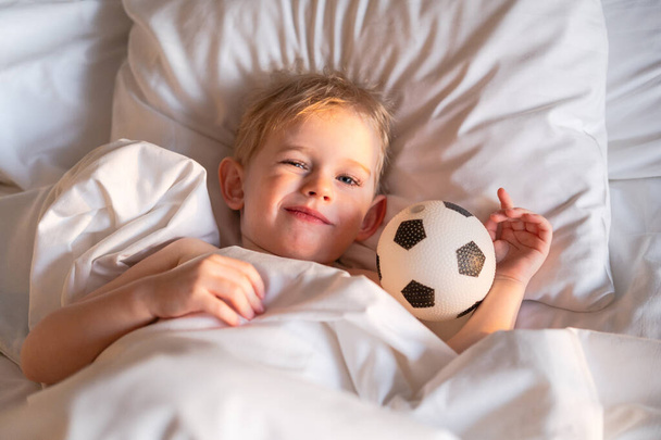 Çocuk uyanıyor ve bir futbol topuyla sarılıp güneşten gözlerini kısıyor.. - Fotoğraf, Görsel