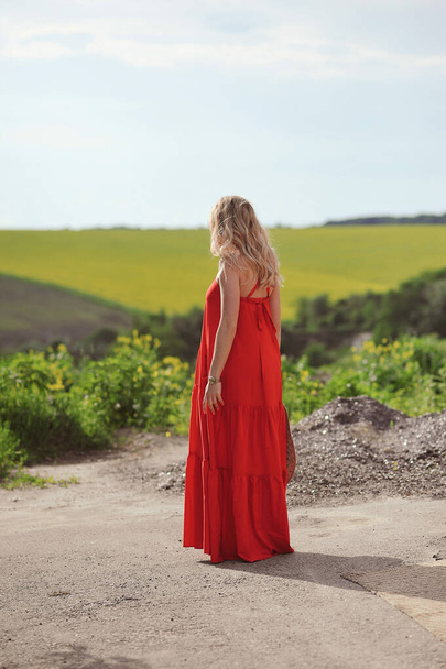 Οπίσθια όψη της ξανθιάς γυναίκας με κόκκινο φόρεμα κοιτάζοντας προς την απόσταση στη φύση και κρατώντας ψάθινο καπέλο - Φωτογραφία, εικόνα