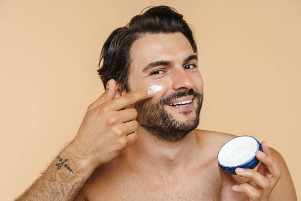 Young half-naked man smiling while applying face cream isolated over beige background - Foto, imagen