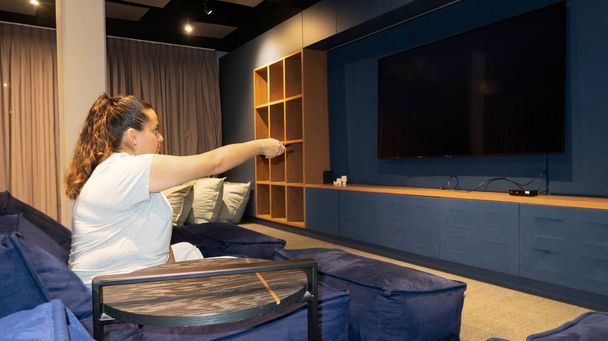 Young girl in big screen home cinema room stretches out hand with remote control - Photo, Image