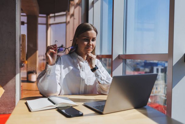 Вид зосередженої жінки в повсякденному вбранні на нетбуці під час роботи над новим проектом, що сидить за столом у сучасному робочому просторі
 - Фото, зображення