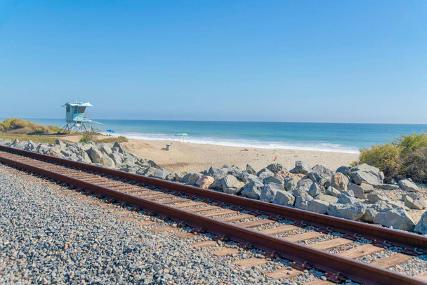 Tren cerca de la playa en San Clemente, California. Hay una casa de salvavidas azul claro en la orilla arenosa contra la vista del cielo y el azul. - Foto, imagen