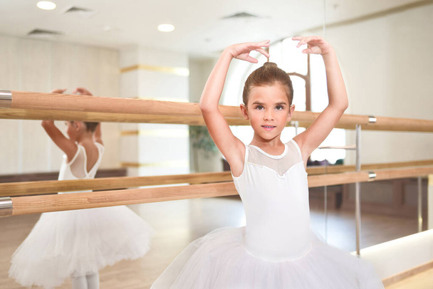 Portrait of a cute little ballerina standing with her hands up in a performance dress. She is dreaming to become a professional ballet dancer. Dance school, ballet studio. - Фото, изображение