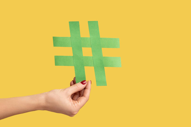 Closeup of woman hand holding large green paper hashtag symbol, hash sign of famous media content, social media marketing and blog promotion. Indoor studio shot isolated on yellow background. - Foto, Imagen