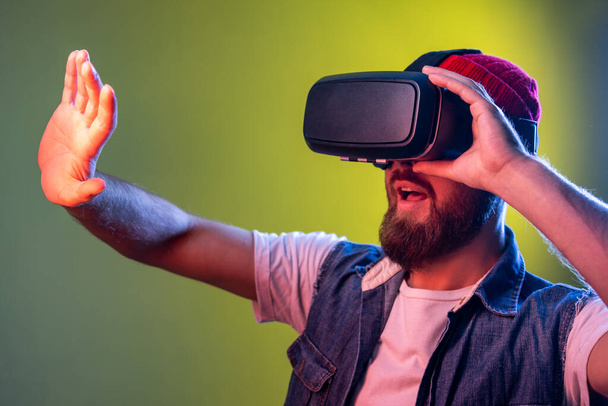 Portrait of scared gamer hipster man with beard in vr glasses, stretching arm forward while playing virtual reality game, frightened face. Indoor studio shot isolated on colorful neon light background - Photo, image