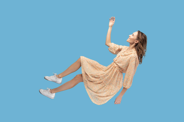 Hovering in air. Cheerful smiling pretty girl in yellow dress levitating flying in mid-air, looking up happy dreamy and raising hand to catch. indoor studio shot isolated on blue background - Photo, image