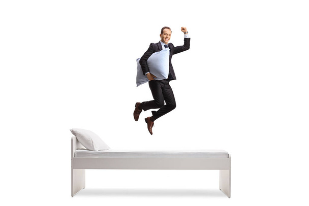 Full length shot of a happy businessman holding a pillow and jumping above bed isolated on white background - Valokuva, kuva
