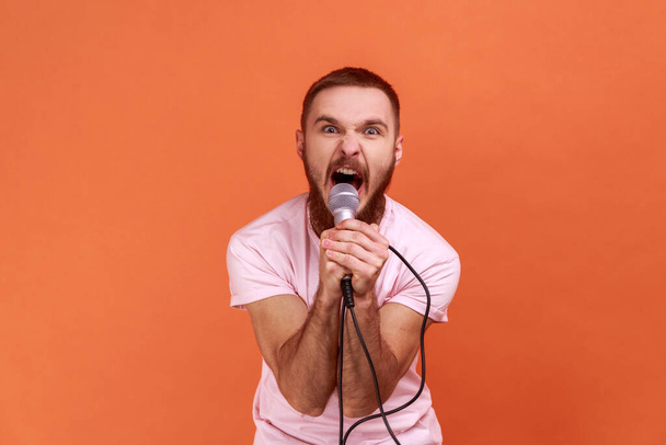 Portrait of angry crazy bearded man holding microphone in hands, looking at camera and screaming loudly, wearing pink T-shirt. Indoor studio shot isolated on orange background. - Foto, immagini