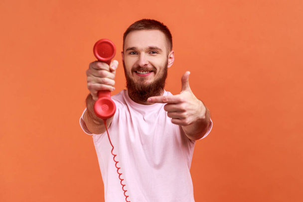 Portrait of smiling happy bearded man pointing finger at retro phone handset to camera, asking to answer phone, wearing pink T-shirt. Indoor studio shot isolated on orange background. - Fotó, kép