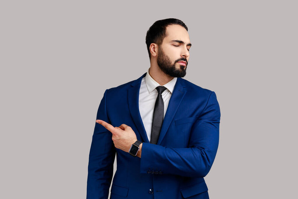 Portrait of bearded man pointing to the side and turning away with angry vexed face, giving order to leave, wearing official style suit. Indoor studio shot isolated on gray background. - Foto, immagini