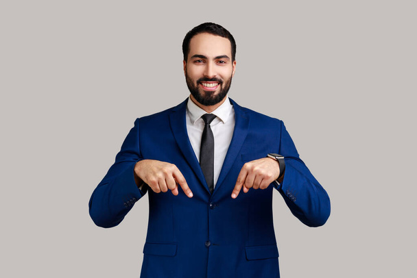 Handsome bearded businessman pointing down place for commercial idea, looking at camera with toothy smile, wearing official style suit. Indoor studio shot isolated on gray background. - Fotoğraf, Görsel