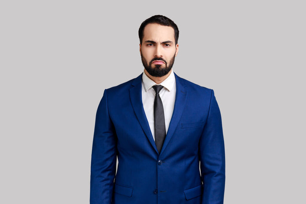 Portrait of sad upset bearded man standing and looking at camera with dissatisfied sadness face, expressing sorrow, wearing official style suit. Indoor studio shot isolated on gray background. - Foto, Imagen