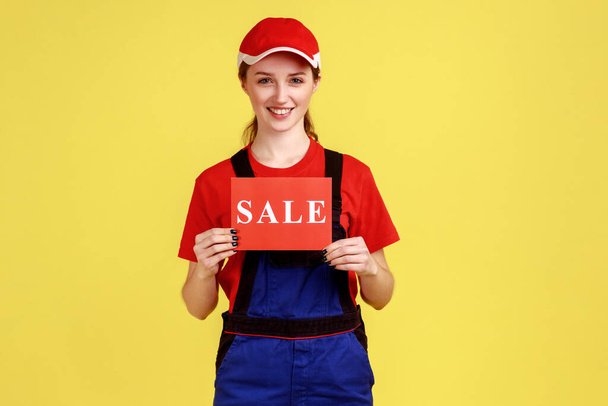 Portrait of positive attractive handy woman standing and holding card with sale inscription, discounts for service, wearing overalls and red cap. Indoor studio shot isolated on yellow background. - Foto, Imagem