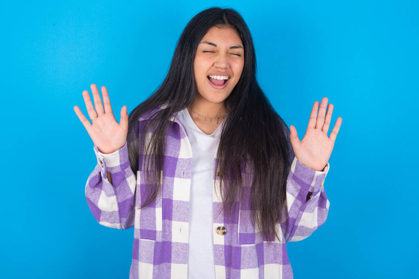 Emotive young latin woman wearing plaid shirt over blue background laughs loudly, hears funny joke or story, raises palms with satisfaction, being overjoyed amused by friend - Foto, Bild
