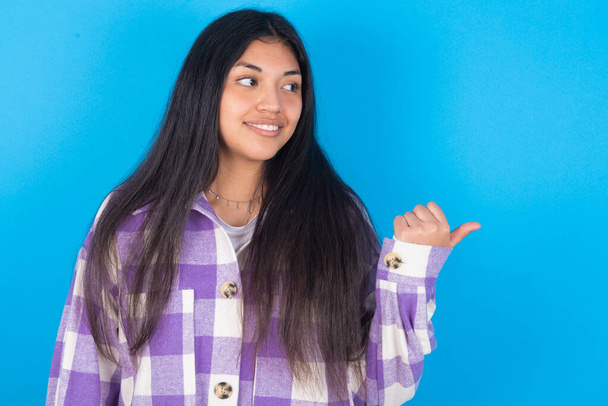 Encantadora jovem latina vestindo camisa xadrez sobre fundo azul olhando para o espaço de cópia com anúncios - Foto, Imagem