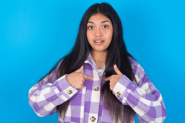 Mulher latina jovem envergonhada vestindo camisa xadrez sobre fundo azul indica em si mesma com expressão confusa, sendo chocada por ser escolhida para participar da competição, hesita sobre algo - Foto, Imagem