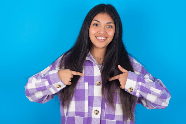 young latin woman wearing plaid shirt over blue background points at his body, being in good mood after going shopping and making successful purchases - Foto, imagen