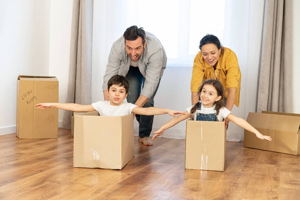 Family moving in new home. Multiracial family of four have fun in new house, parents riding kids in cardboard boxes in empty living room, happy children spreading arms and laughing - Photo, image