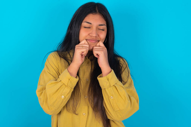 Pleased young latin woman wearing yellow shirt over blue background with closed eyes keeps hands near cheeks and smiles tenderly imagines something very pleasant - Fotó, kép