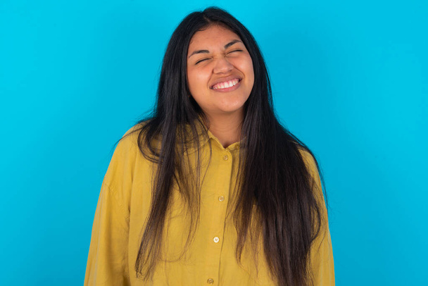Позитивная молодая латинская женщина в жёлтой рубашке на синем фоне с восторженным выражением лица закрывает глаза и смеется показывает белые идеальные зубы - Фото, изображение