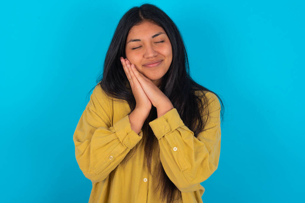 young latin woman wearing yellow shirt over blue background leans on pressed palms closes eyes and has pleasant smile dreams about something - Zdjęcie, obraz