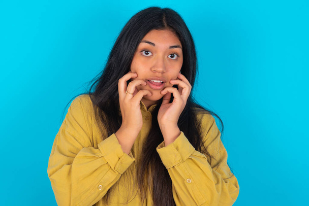 Speechless young latin woman wearing yellow shirt over blue background keeps hands near opened mouth reacts to shocking news stares wondered at camera - Foto, imagen