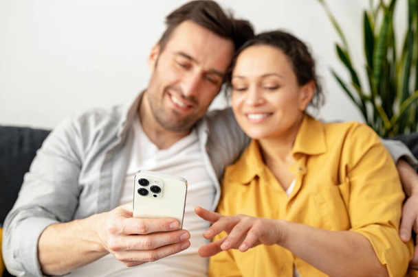 Emocionada y muy alegre pareja caucásica sentada en el sofá en casa y sosteniendo el teléfono inteligente. Hombre y mujer viendo la película - Foto, imagen