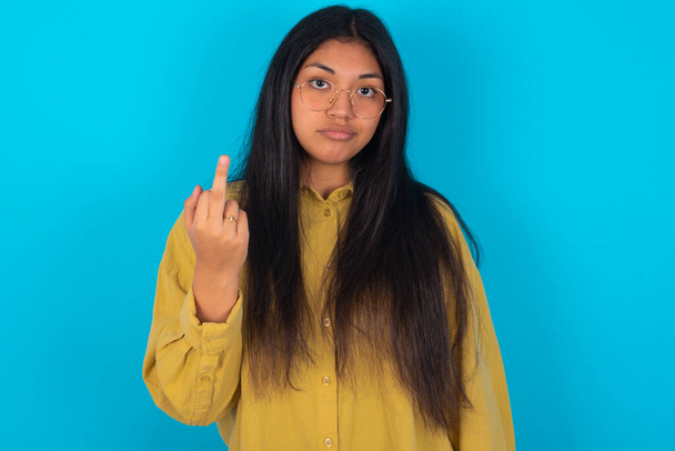 молодая латинская женщина в желтой рубашке на синем фоне показывает средний палец плохой знак просит не беспокоиться. Провокация и грубое отношение. - Фото, изображение