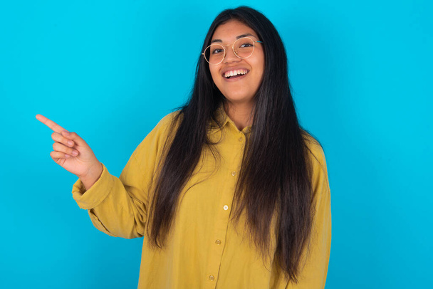 young latin woman wearing yellow shirt over blue background laughs happily points away on blank space demonstrates shopping discount offer, excited by good news or unexpected sale. - Fotografie, Obrázek