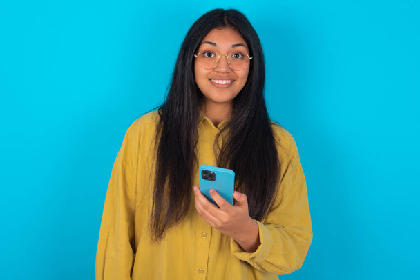 young latin woman wearing yellow shirt over blue background holds mobile phone in hands and rejoices positive news, uses modern cellular - Zdjęcie, obraz